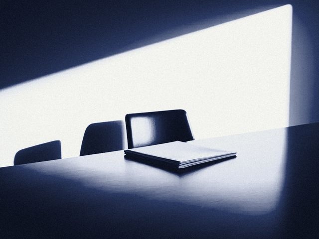 An empty conference table with a closed notebook, in hard window light.